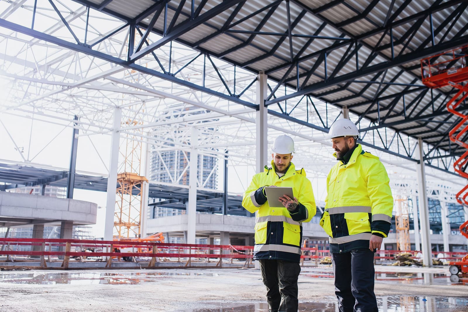 Two contractors in hi-vis reviewing progress on a tablet inside a steel-framed facility under construction