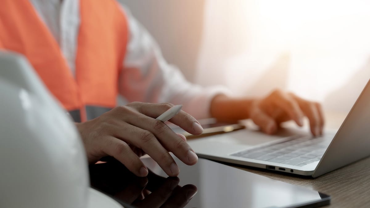 Close-up of a project engineer working on a laptop with a hard hat on the desk beside them