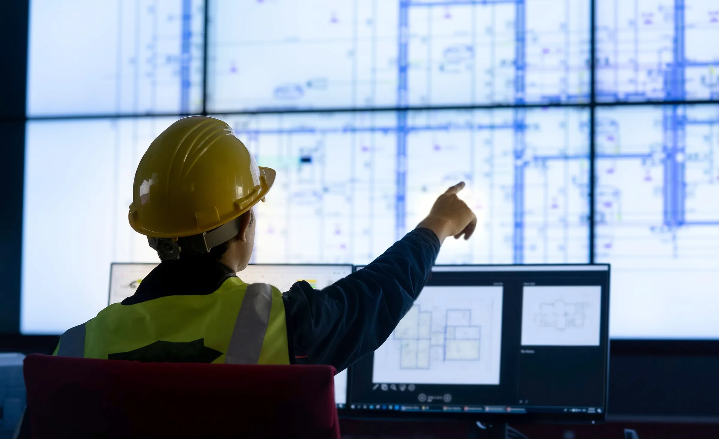 Engineer pointing at a control-room wall of drawings and dashboards, reading project data at a glance