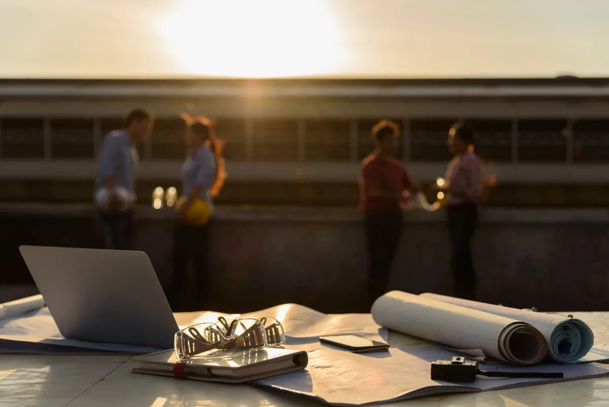 Laptop and rolled drawings on a rooftop table as a project team meets at sunset