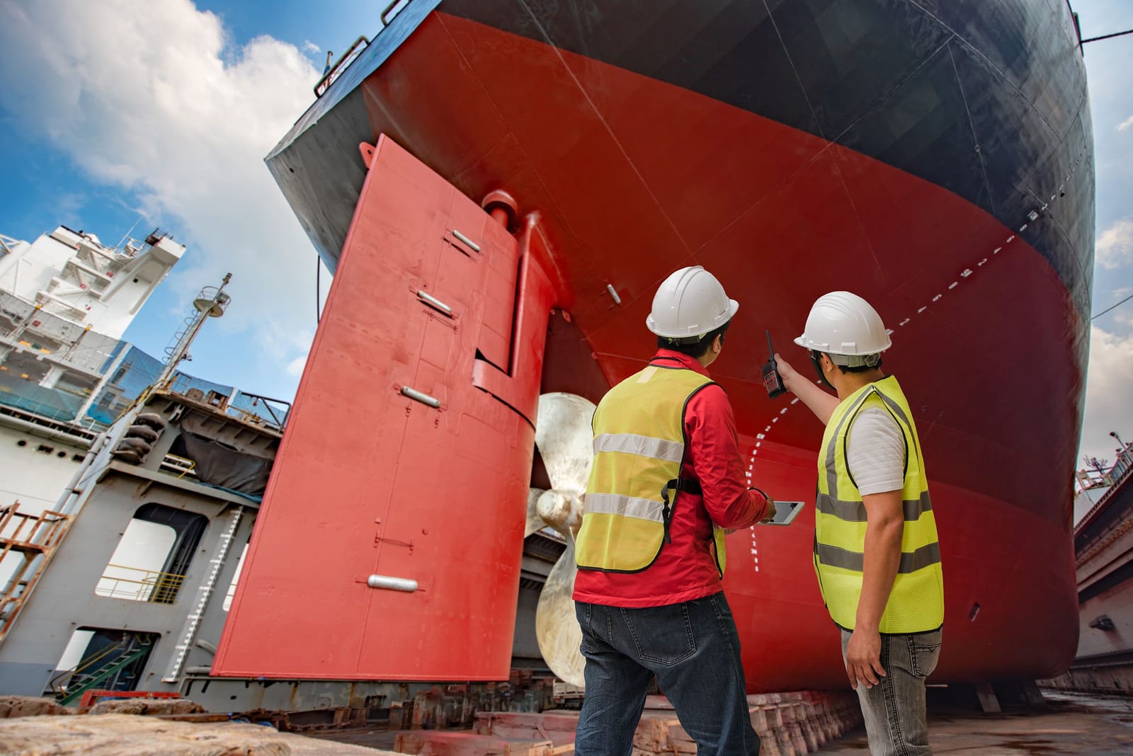 Two shipbuilders inspecting a vessel's red hull in dry dock, recording their checks as they go