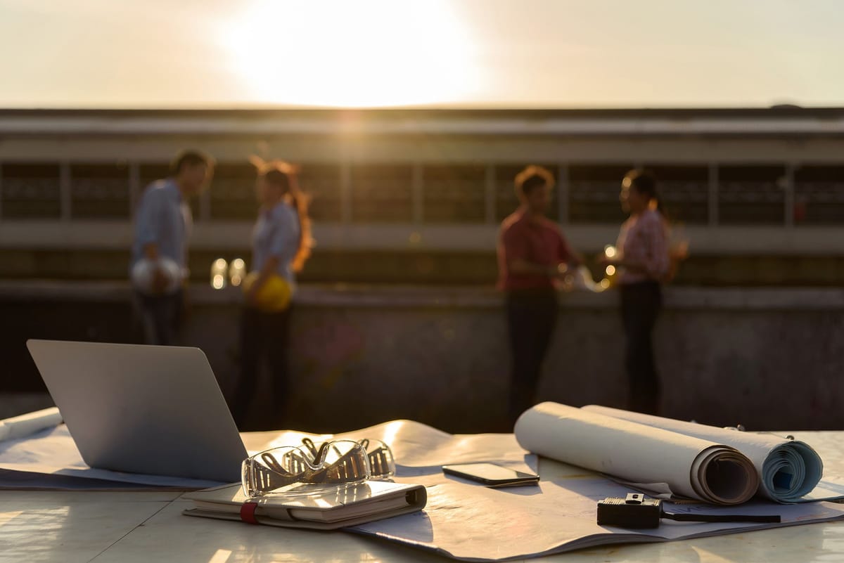 Laptop and rolled drawings on a site table at sunset while the team meets in the background